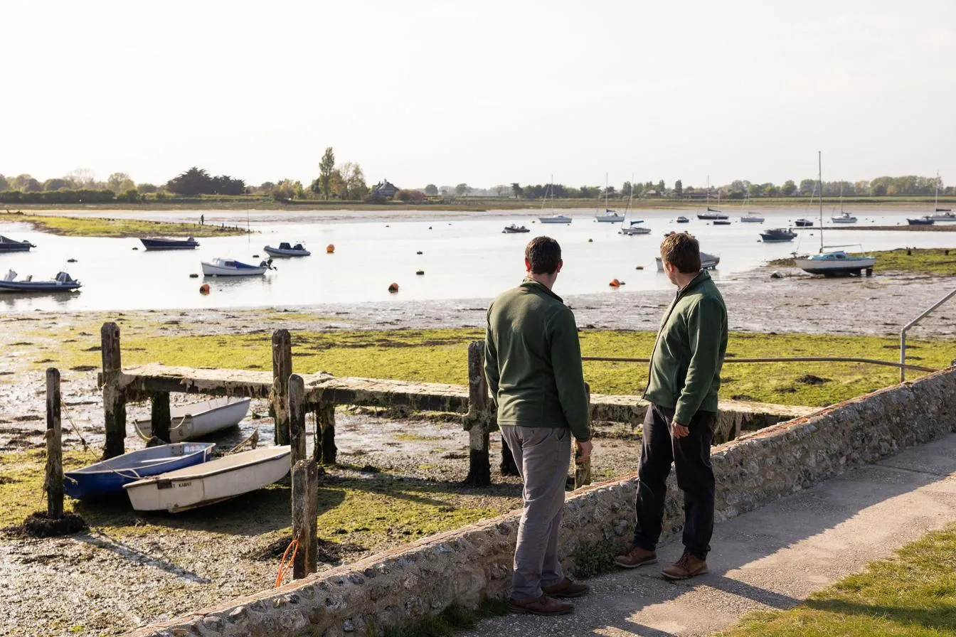 Two people looking out over a tidal harbour at low water, small boats moored beyond the saltmarsh.
